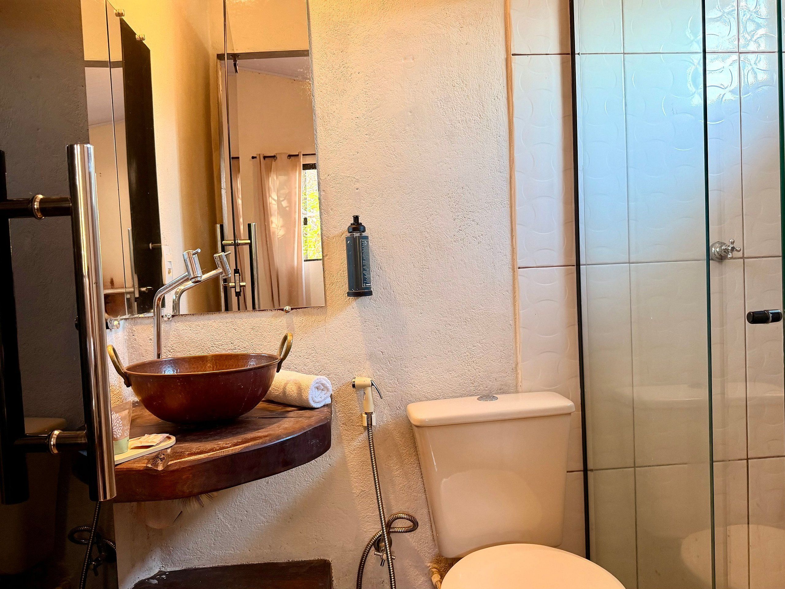 Un baño con tocador de madera, un espejo grande, un lavabo con cuenca de cobre, un inodoro y una zona de ducha.