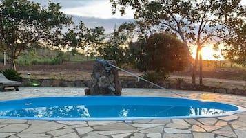 A swimming pool with a stone border, surrounded by trees and a sunset in the background.