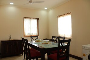 A dining area with a glass table, four chairs, a wooden cabinet, and two windows with curtains. - OYO 9625 Home 2BHK Goan Paradise Colva (Betalbatim)