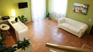 Uma sala de estar com um sofá branco, uma escrivaninha com um laptop, uma cadeira e uma televisão na parede.