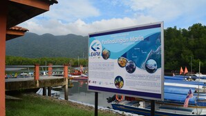 Een bord met informatie over Perindukatan Marin Langkawi, met uitzicht op de kust, boten en een bergachtige achtergrond.