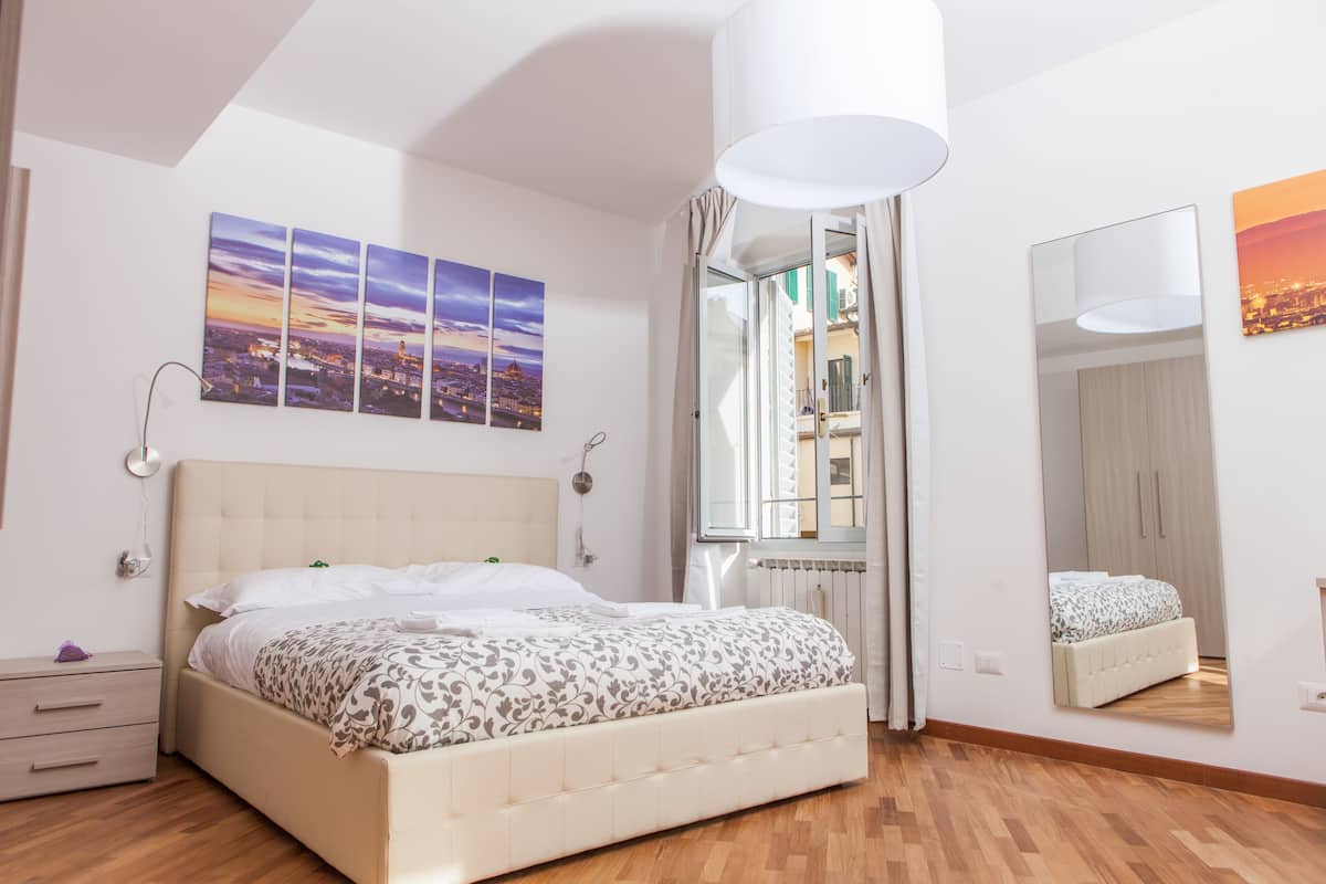 Ein Schlafzimmer mit einem großen Bett, einem Nachttisch, einem Spiegel und Blick auf die Stadt durch ein Fenster.