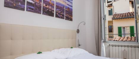 Una habitación de hotel con una cama, un ventanal con vistas a edificios y una pared con tres cuadros.