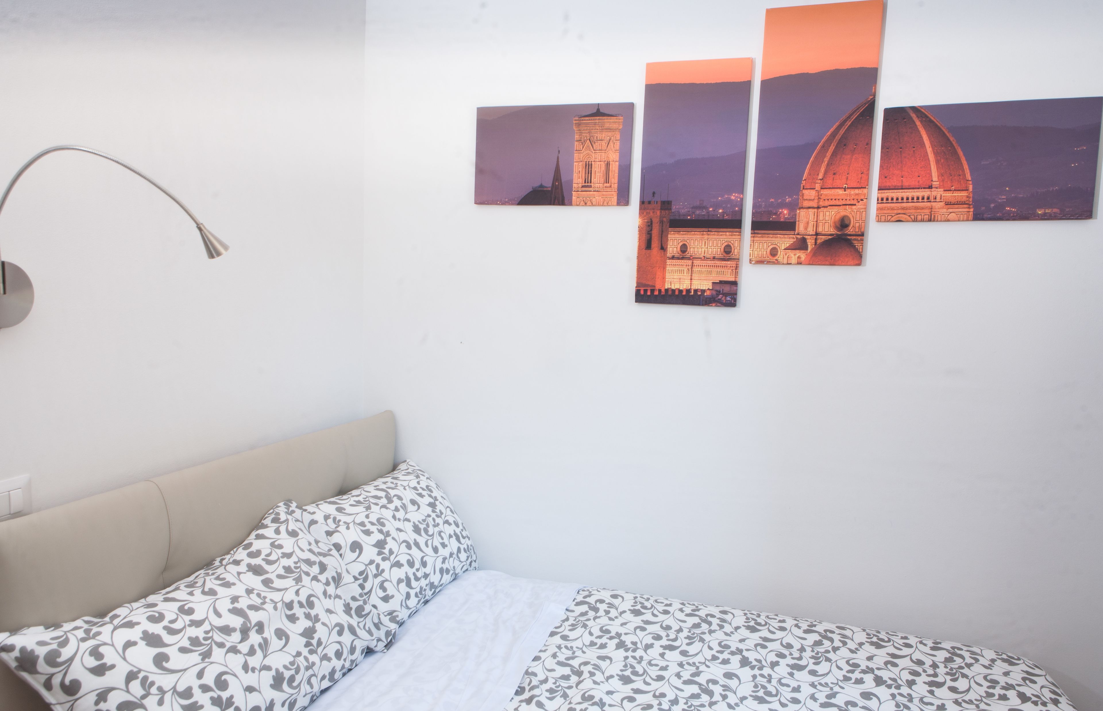A bedroom with a bed, a wall-mounted lamp, and three framed pictures of a building.