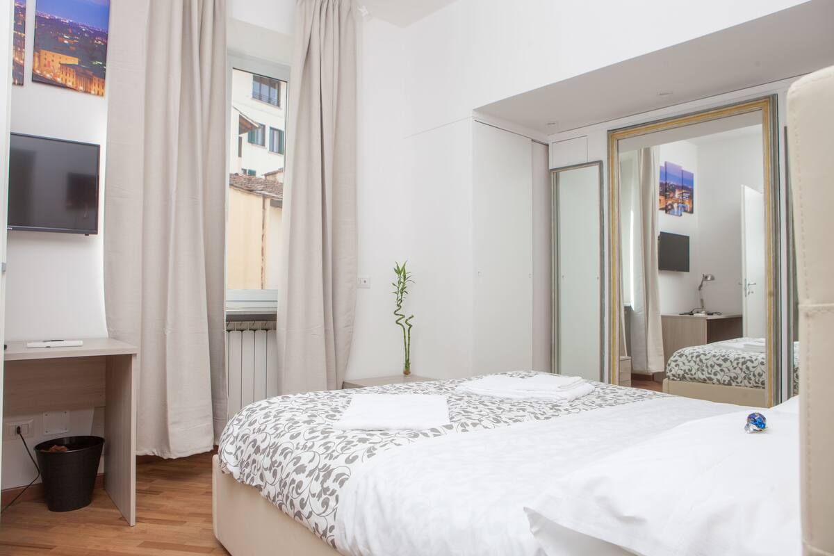 Ein Hotelzimmer mit Bett, Schreibtisch, Fernseher, einem Fenster mit Vorhängen, einem Spiegel und einer Pflanze.