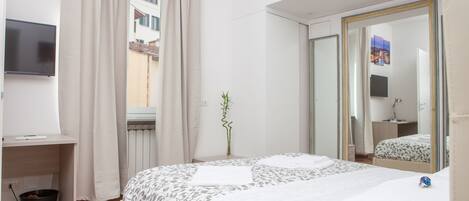 A hotel room with a bed, a desk, a TV, a window with curtains, a mirror, and a plant.