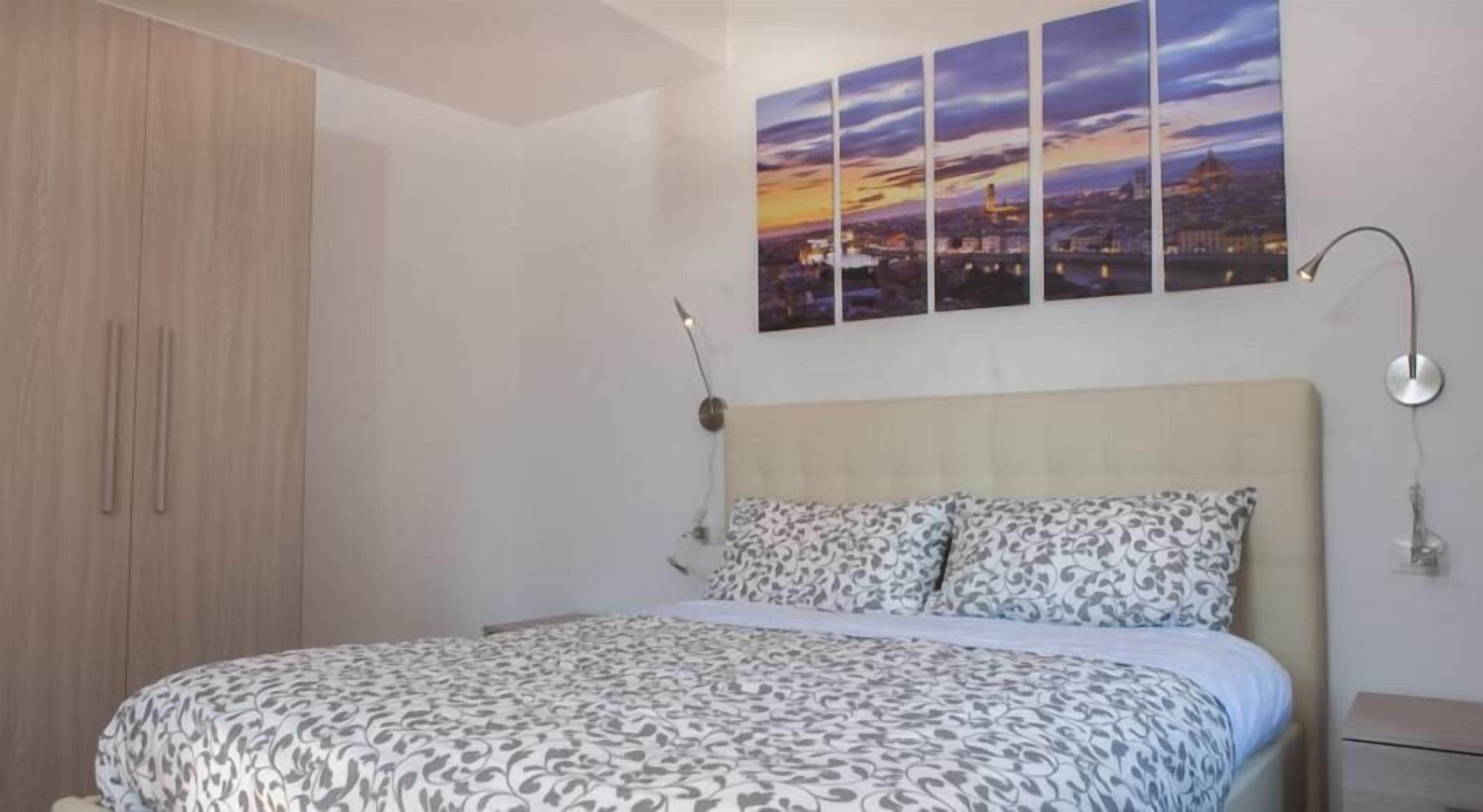 Een slaapkamer met een bed, nachtkastje, lamp, garderobe en een schilderij van een stadsgezicht aan de muur.