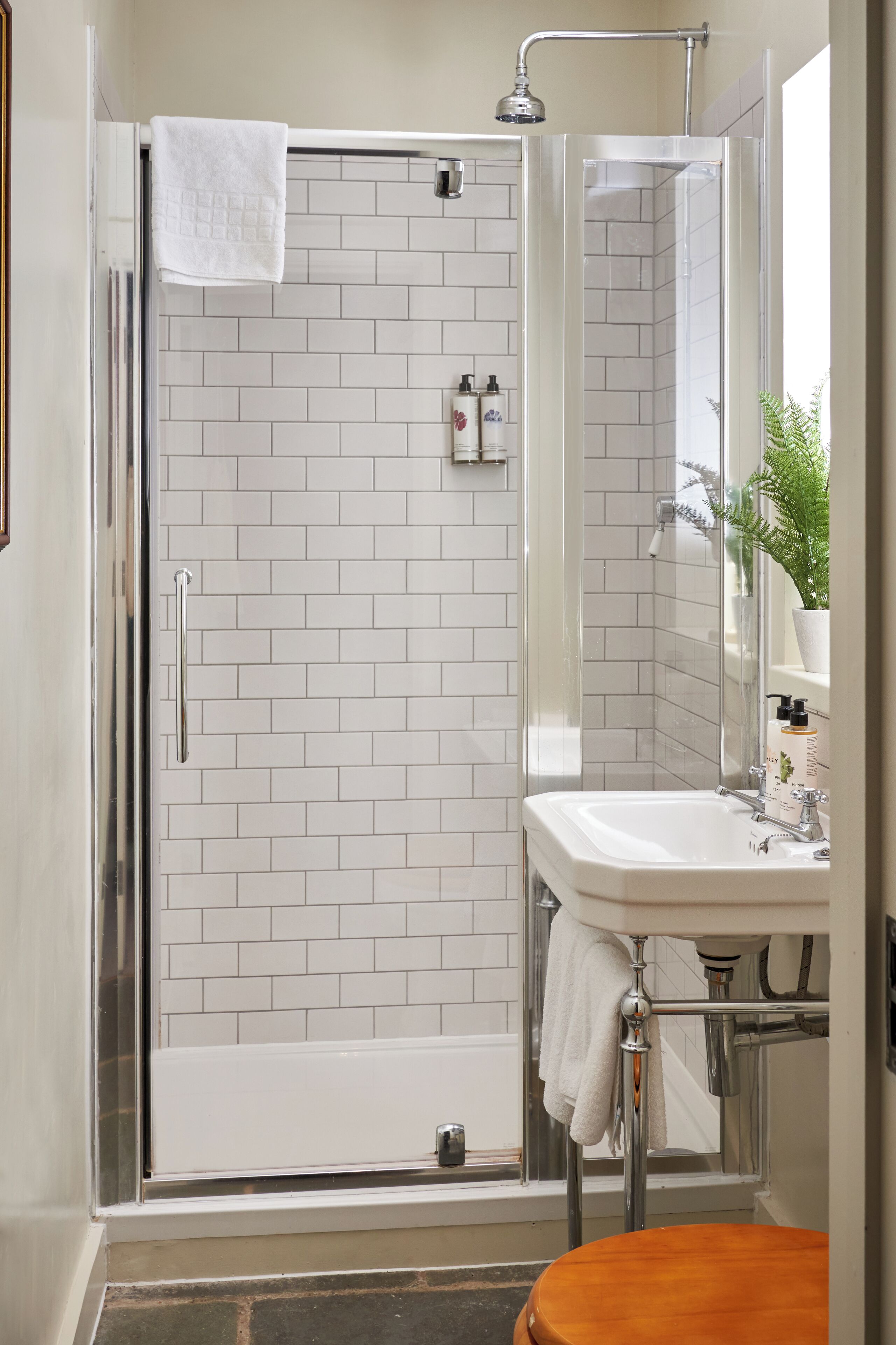 A bathroom with a glass-enclosed shower, white tiled walls, a white sink, and a wooden toilet.