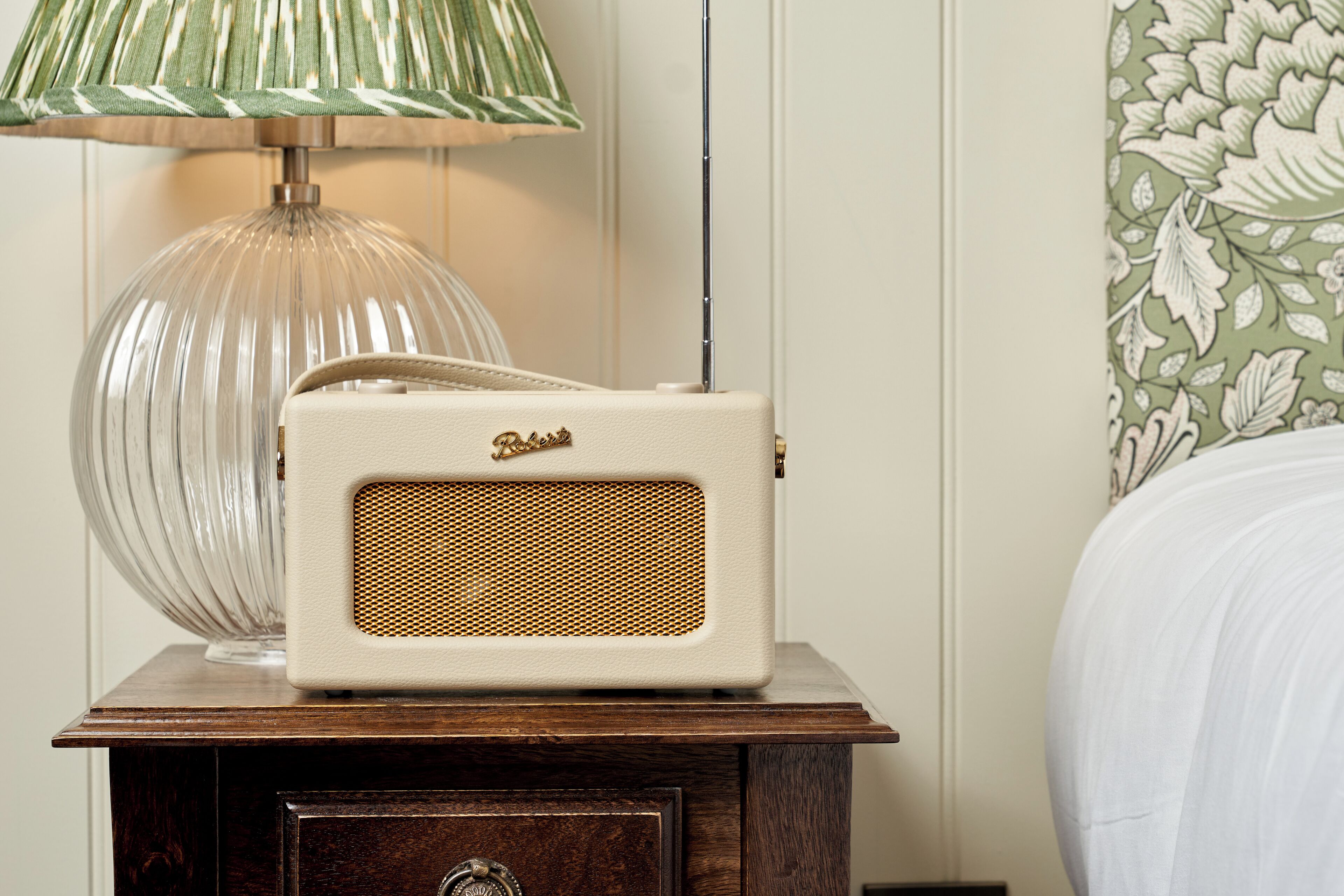A vintage radio on a wooden nightstand with a lamp and a floral wallpaper background.