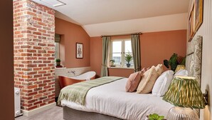 A bedroom with a brick wall, a large bed, a window with curtains, a bedside table with a lamp, and a potted plant. - The Welldiggers Arms (Petworth)
