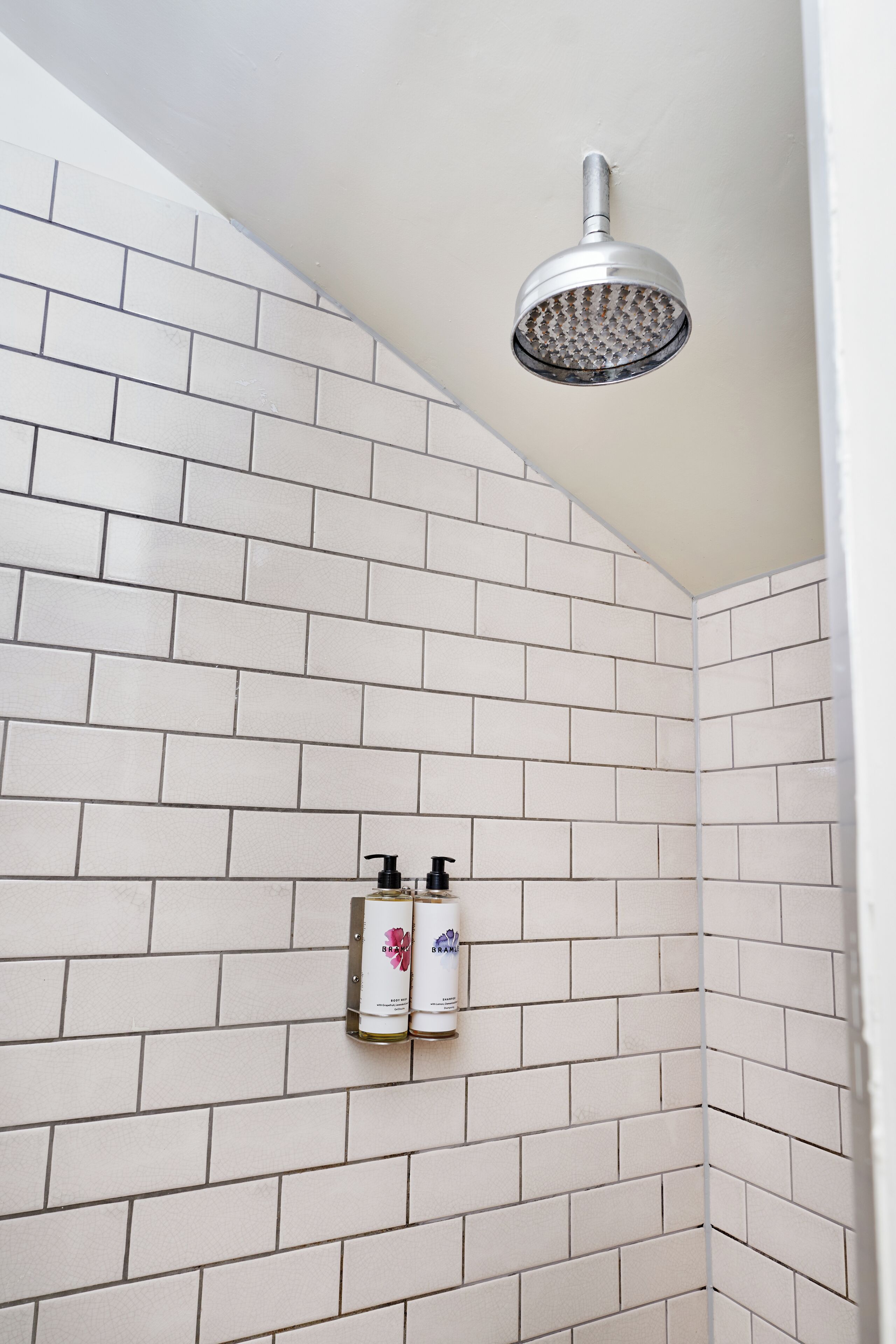 A shower with a handheld showerhead mounted on the wall, white tiled walls, and two bottles of shampoo and conditioner on a small shelf.