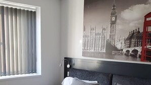 A bedroom with a large bed, a window with blinds, a headboard, and a wall-mounted artwork of a cityscape. - The Bell Gate House (Leicester)
