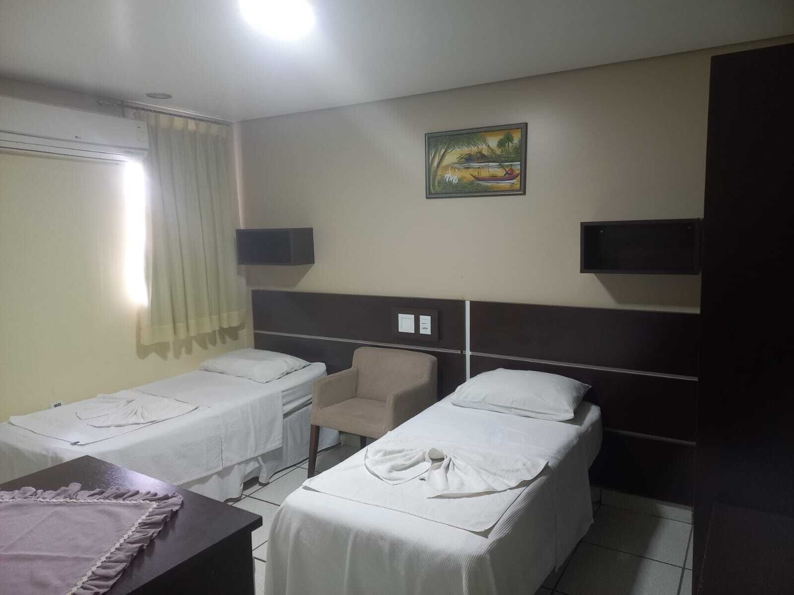 A hotel room with two single beds, a chair, a small table, and a painting on the wall.
