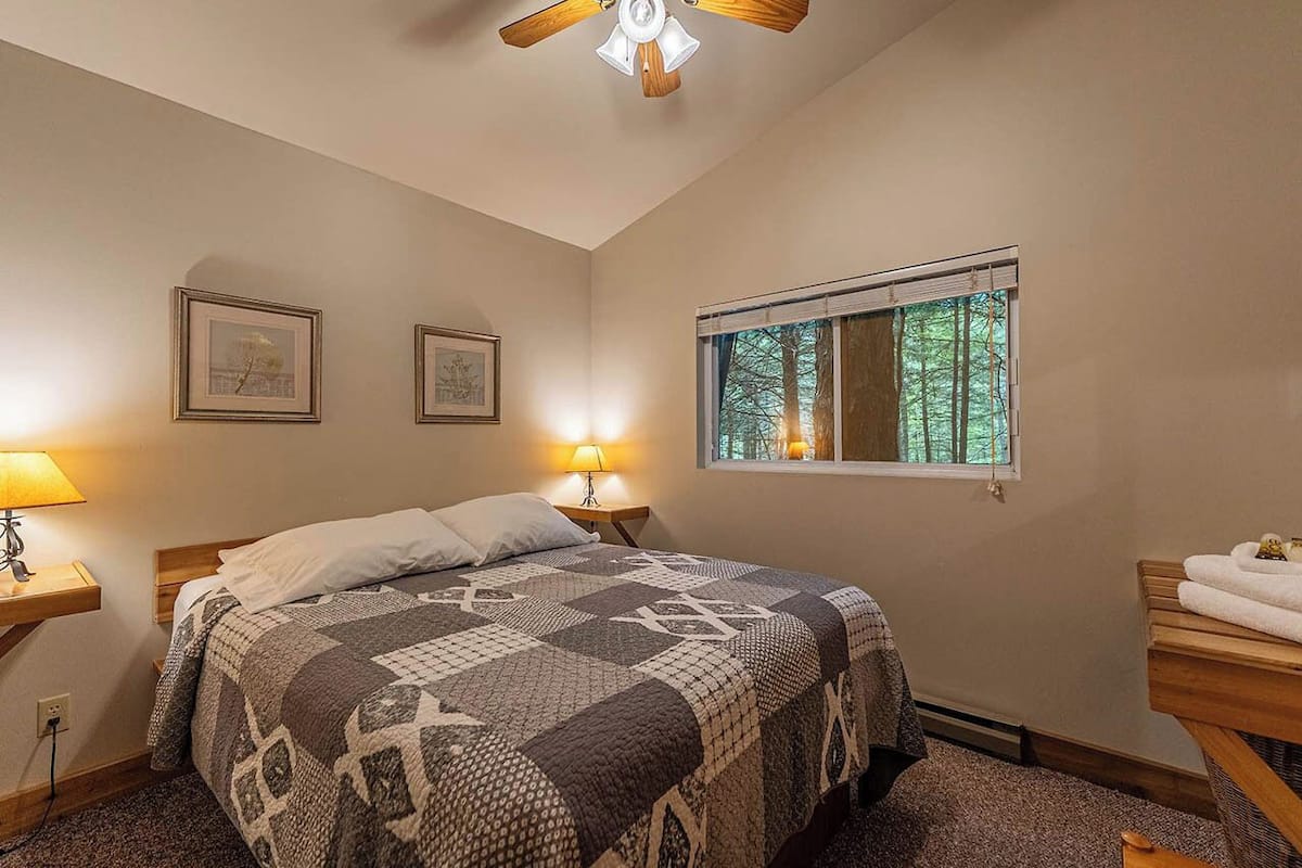 A bedroom with a bed, two bedside lamps, a window with a view of trees, and a ceiling fan.