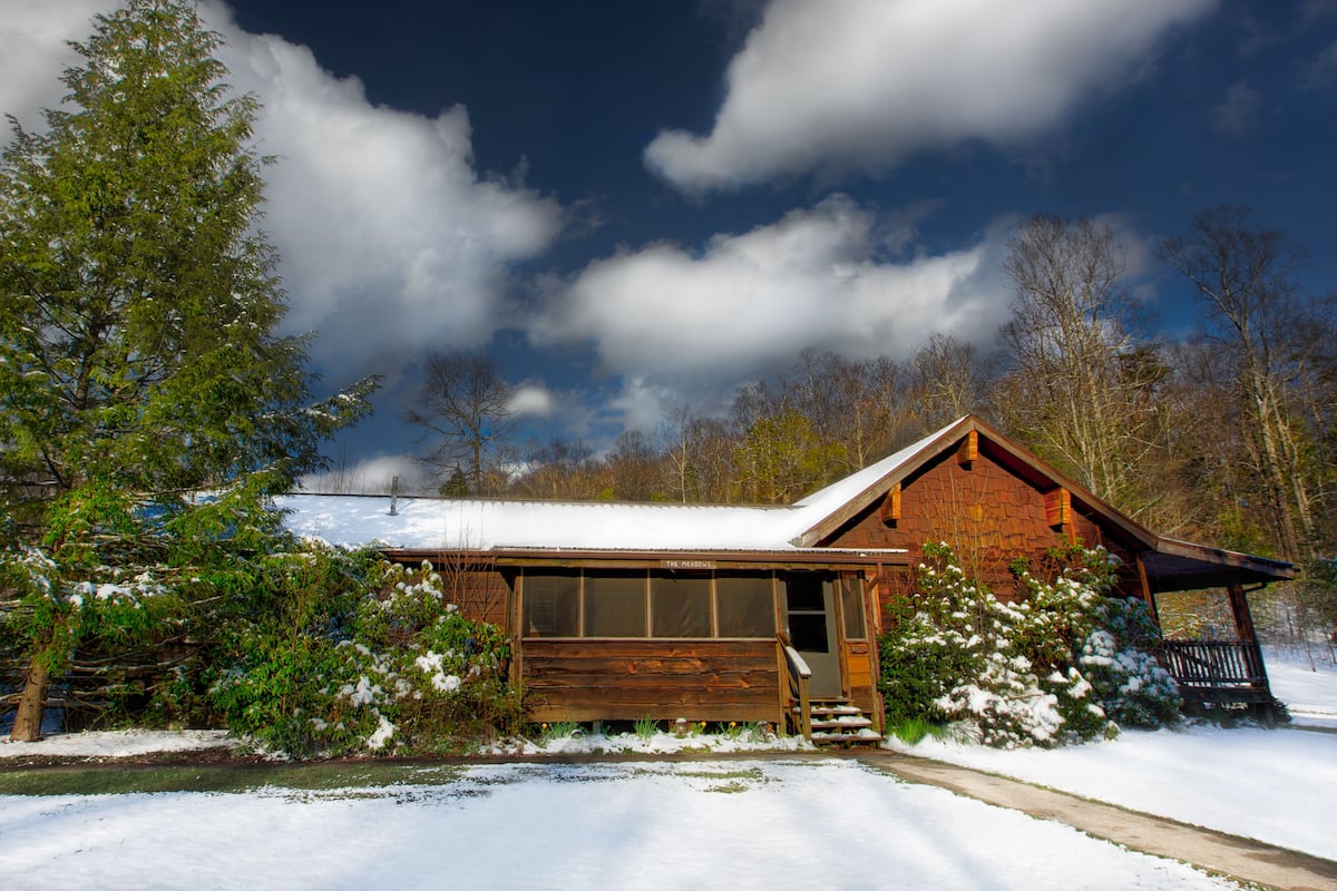 A wooden cabin with a snow-covered roof, surrounded by trees and a clear sky.