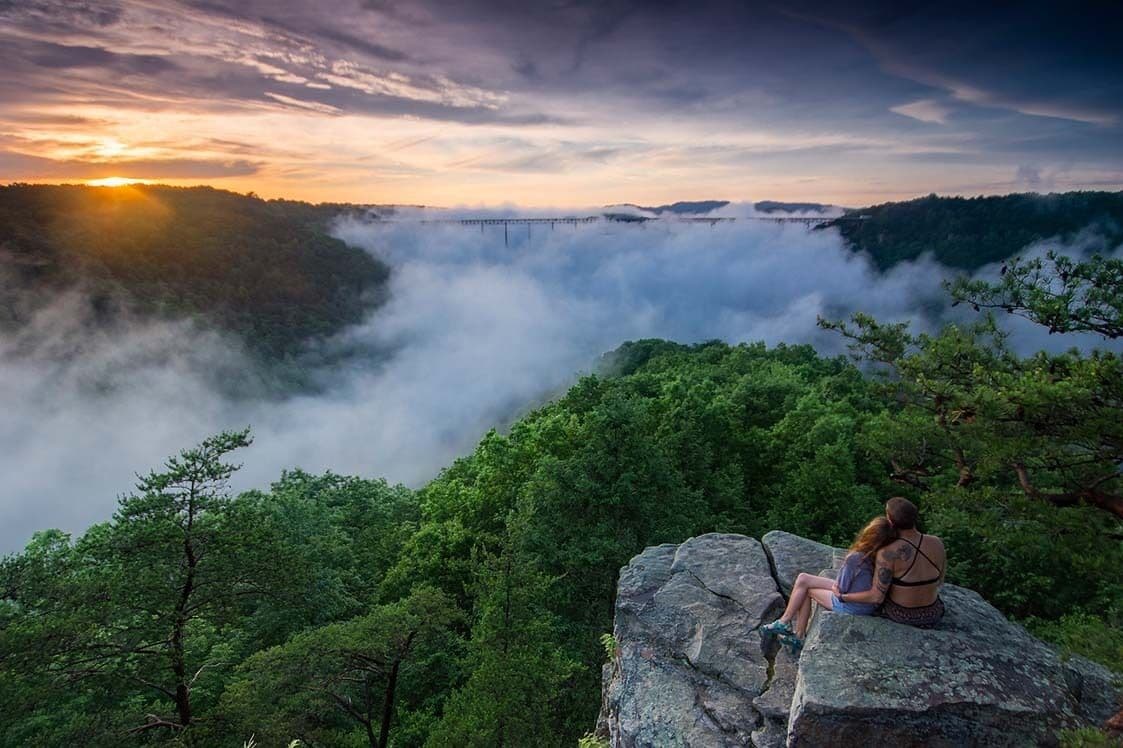 A couple sitting on a rock overlooking a forested valley with a bridge in the distance.