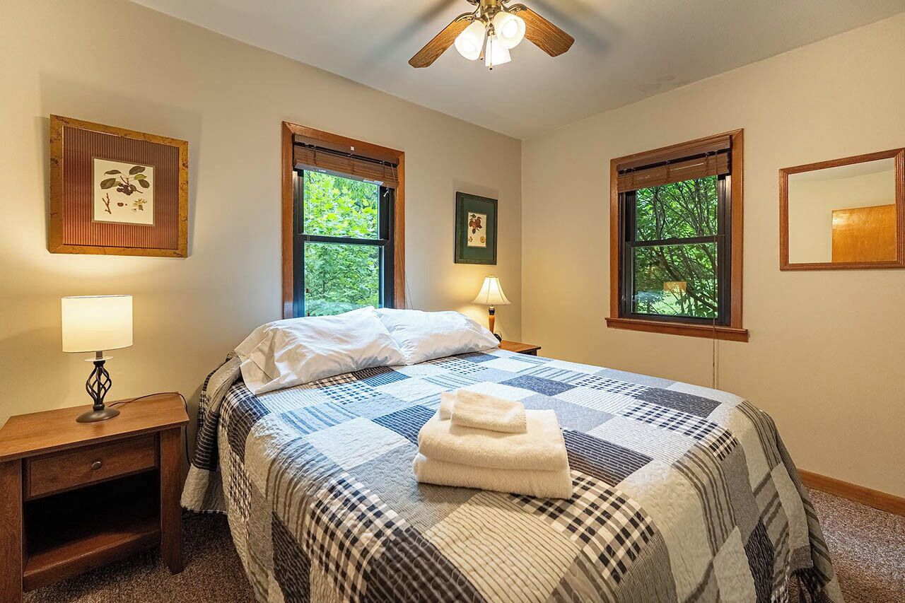 A bedroom with a large bed, two windows, a ceiling fan, and framed pictures on the walls.