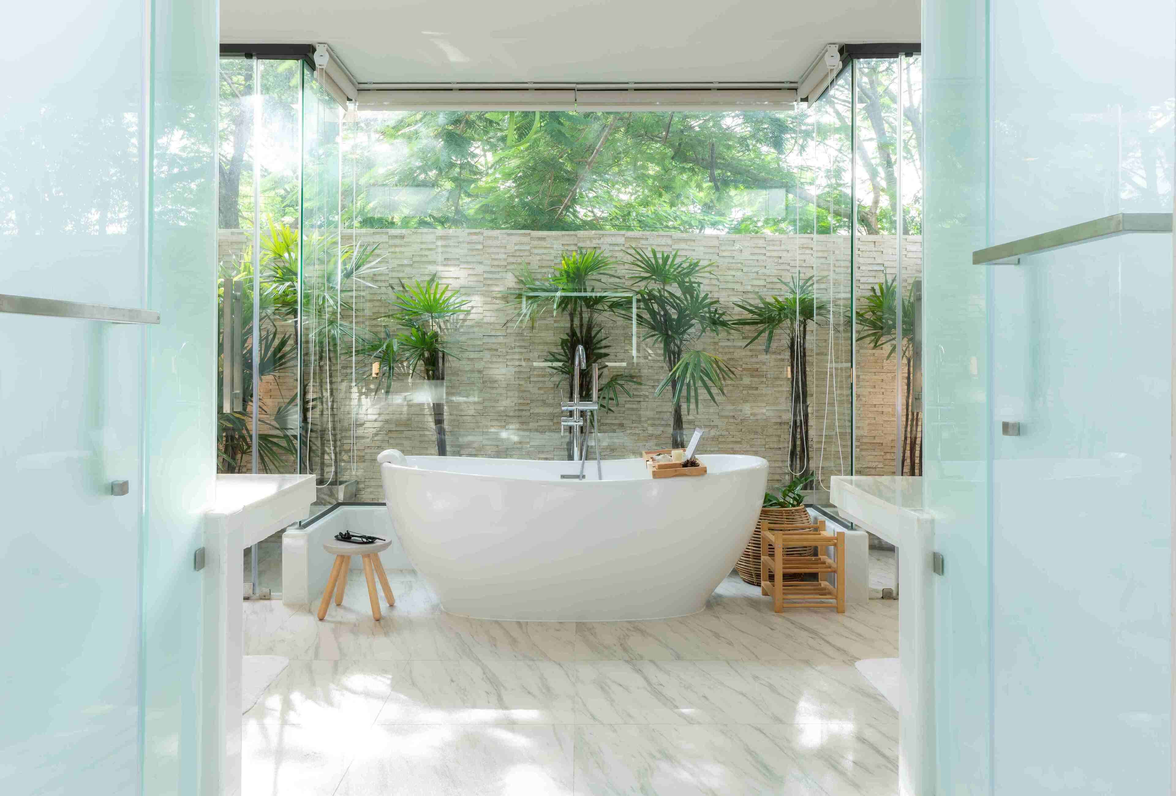 A modern bathroom with a freestanding bathtub, glass walls, and a view of greenery outside.