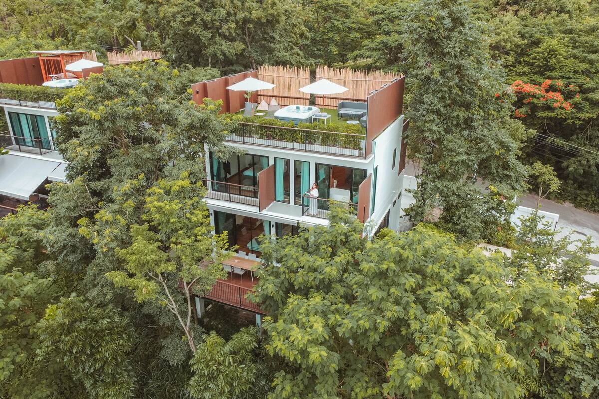 Un edificio moderno de dos pisos con un balcón, rodeado de abundante vegetación.