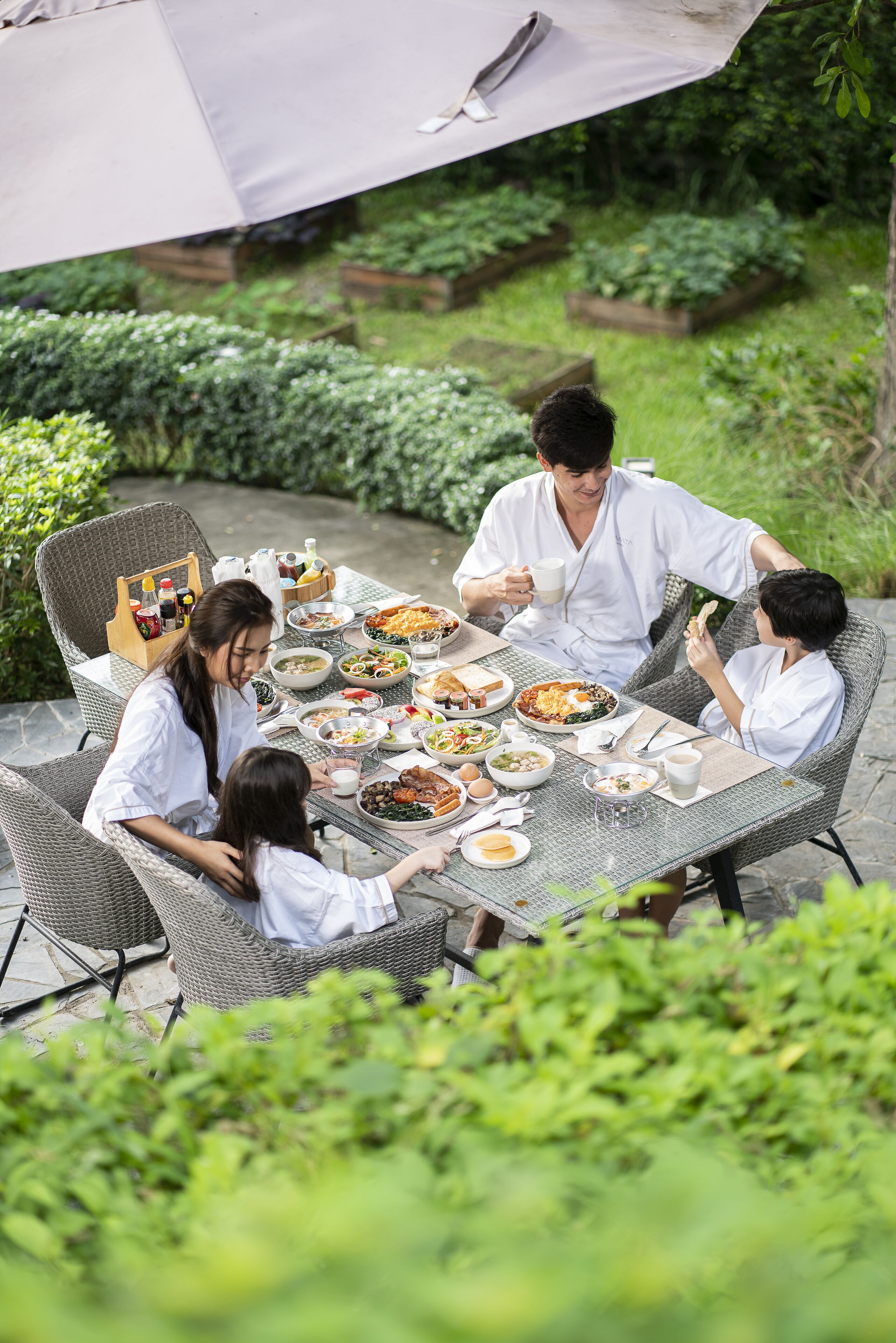 Una familia cenando al aire libre en una mesa con diversos platos, rodeada de sillas de mimbre y una sombrilla.