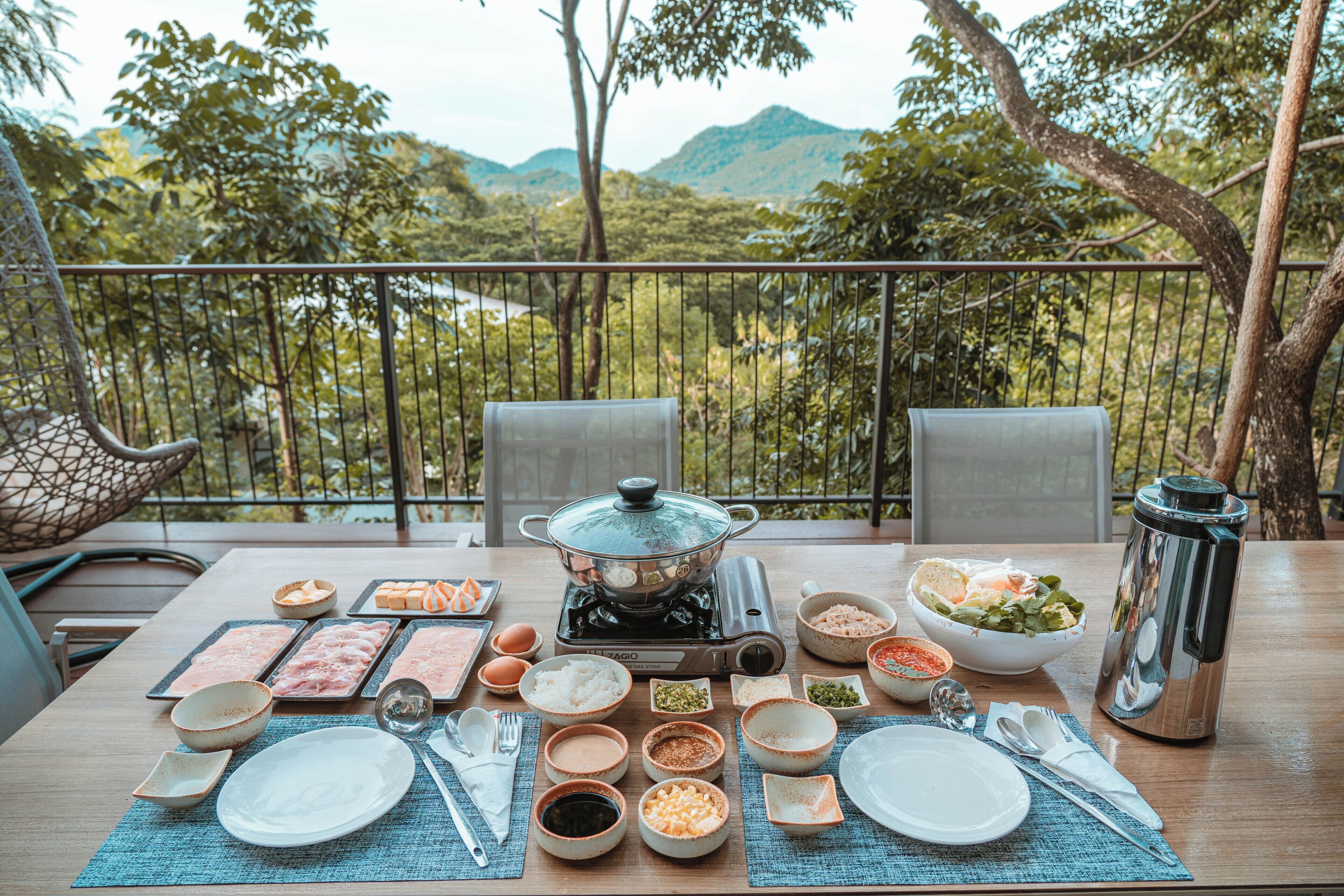 Une table dressée pour un repas, avec différents plats et accompagnements, un réchaud portable et une vue sur un paysage montagneux.
