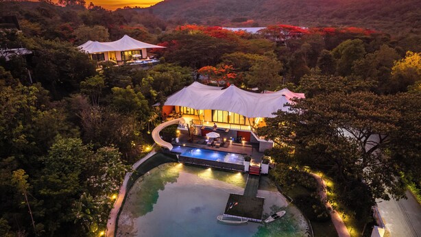 Vista aérea de un resort con piscina, zona de descanso y una gran estructura cubierta, rodeada de abundante vegetación.