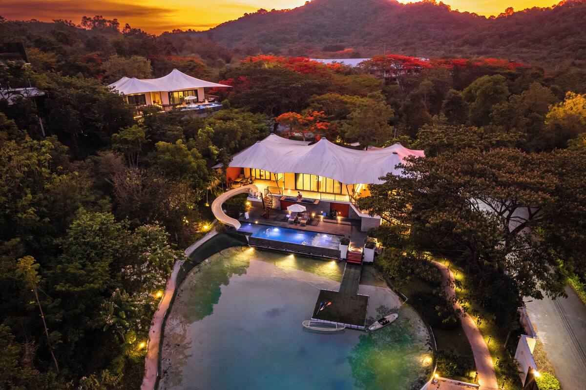 Vista aérea de un resort con piscina, zona de descanso y una gran estructura cubierta, rodeada de abundante vegetación.