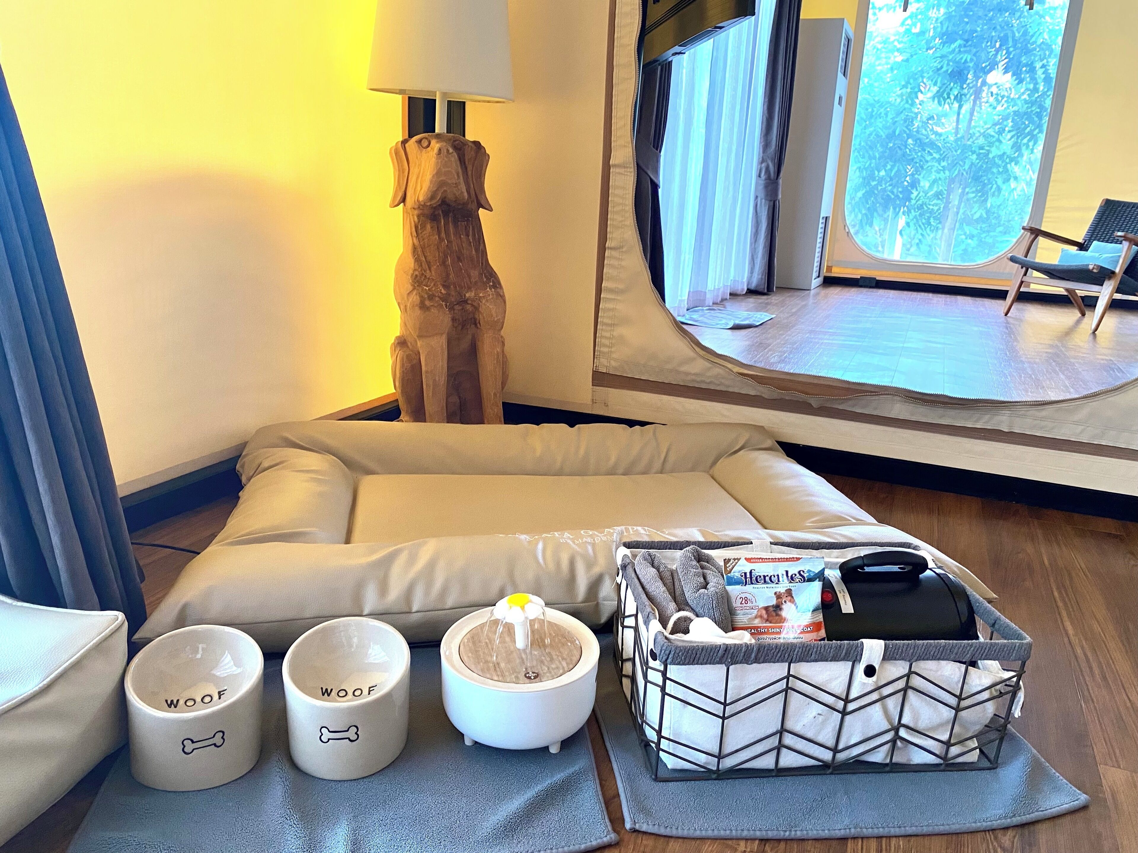 Habitación de hotel con una cama, una escultura de perro de madera, dos tazas con motivos caninos, una fuente de agua y una cesta con alimento para perros.