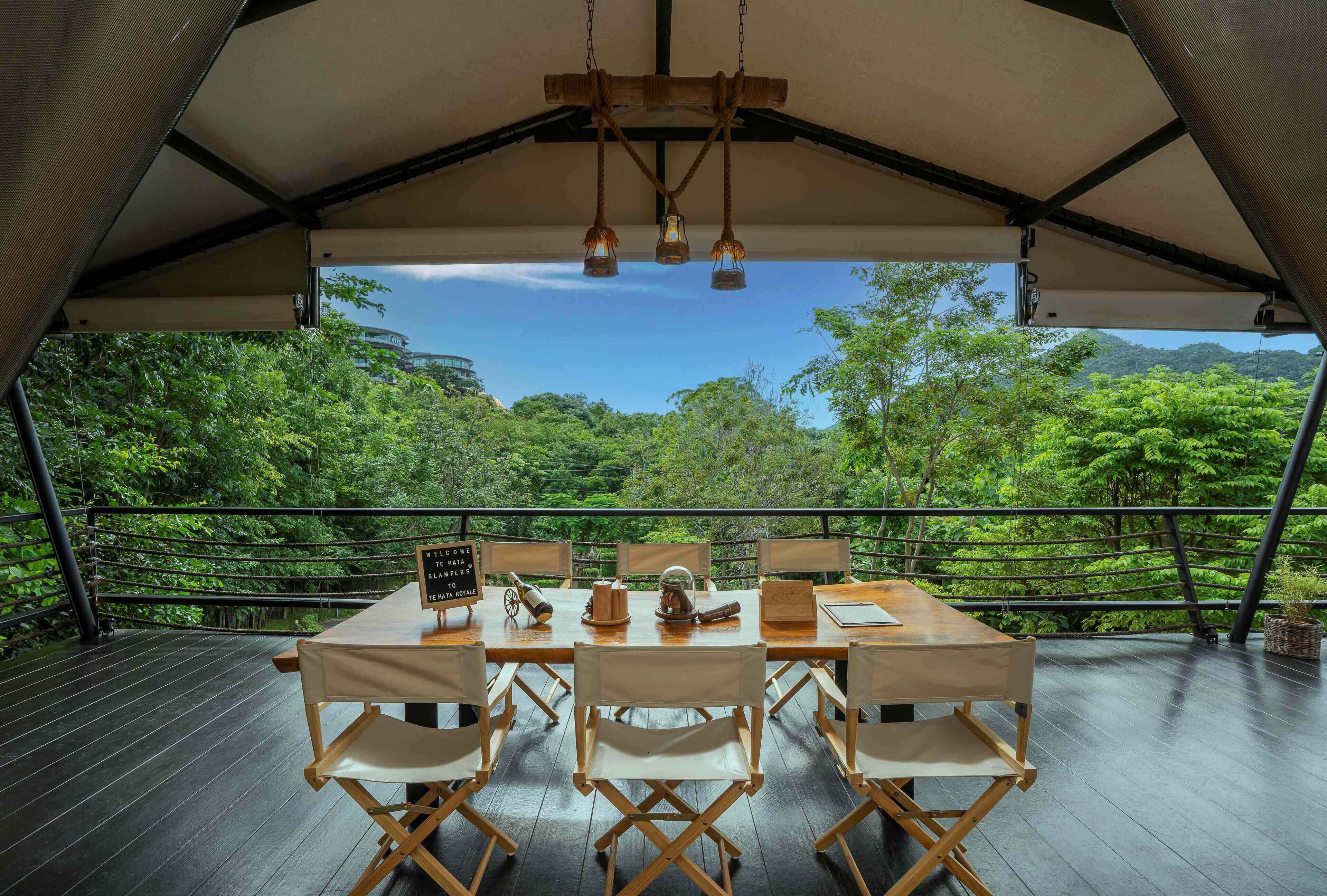 Un área de comedor al aire libre con una mesa dispuesta para seis personas, rodeada por una terraza de madera y vistas a exuberante vegetación y colinas.