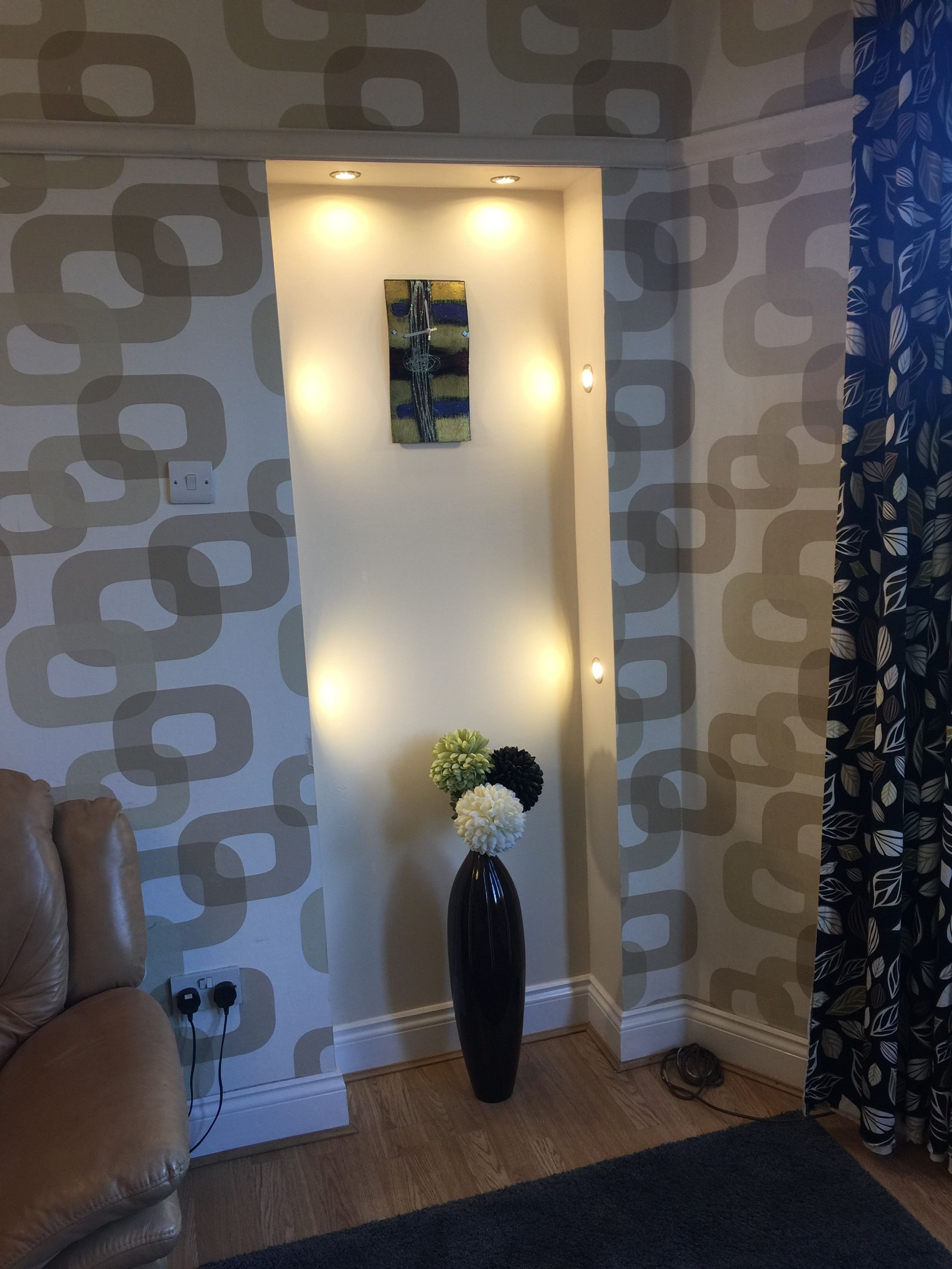 A room with patterned wallpaper, a black vase with flowers, a painting, and a light switch on the wall.