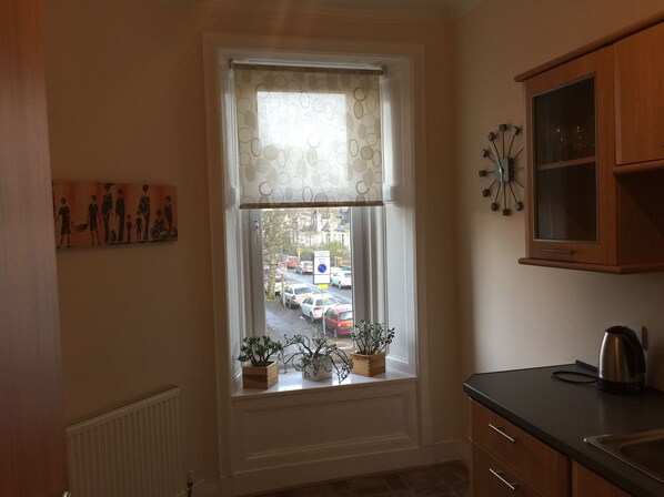 A kitchen with a window, wooden cabinets, a kettle, and a clock on the wall.