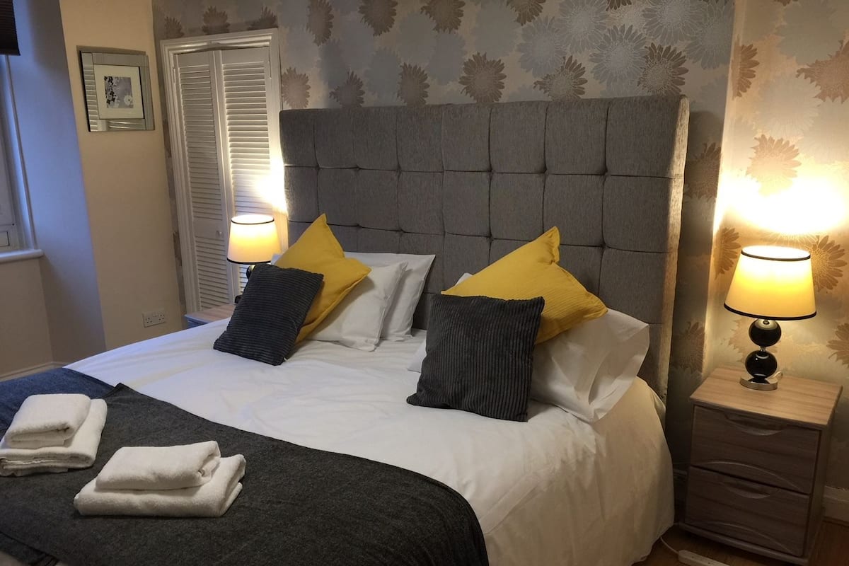 Una cama bien tendida con ropa de cama blanca y gris, dos lámparas de noche y una mesita de noche de madera con cajón.