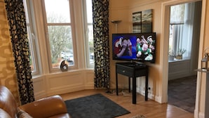 A living room with a leather sofa, a TV on a stand, a window with curtains, and a view of the street outside. - Great location 1 Bedroom Scotstoun Flat (Glasgow)