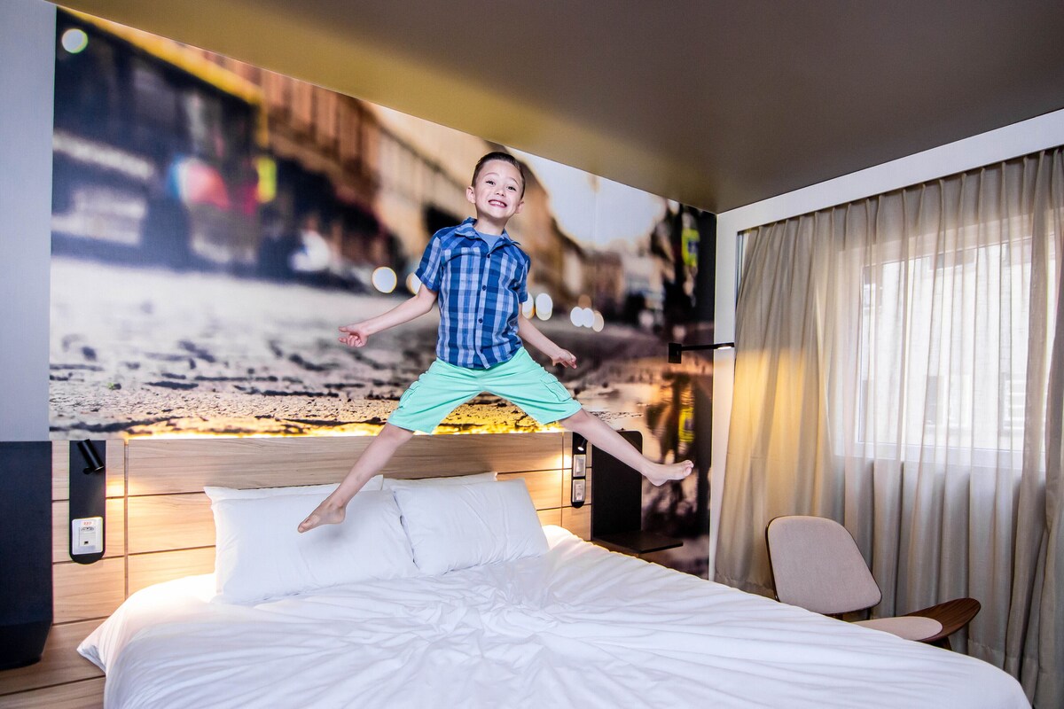 A hotel room with a bed, a nightstand, a chair, and a large wall mural.