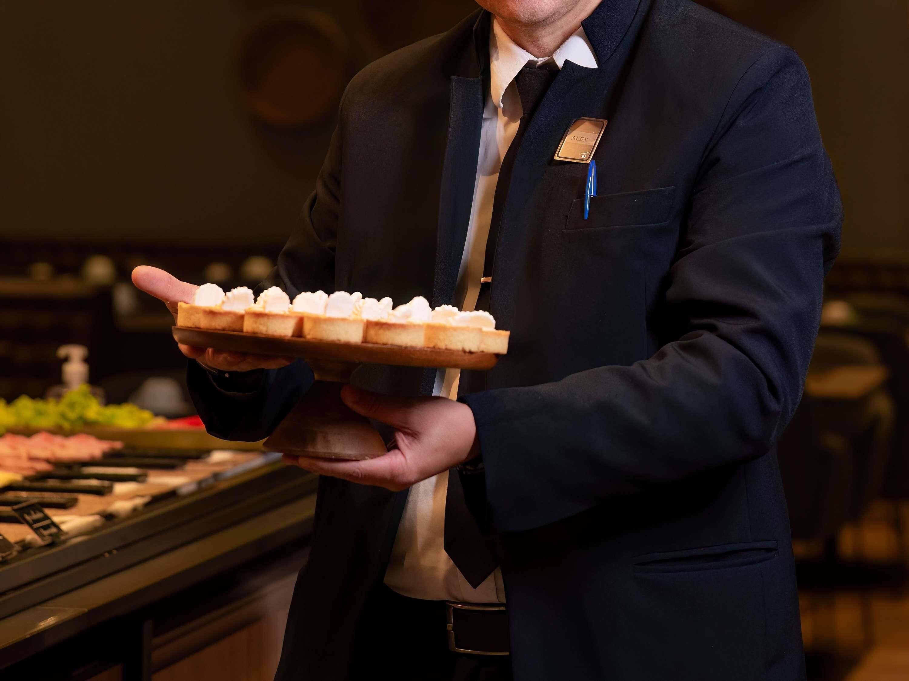 A person in a dark suit holding a tray with small pastries.