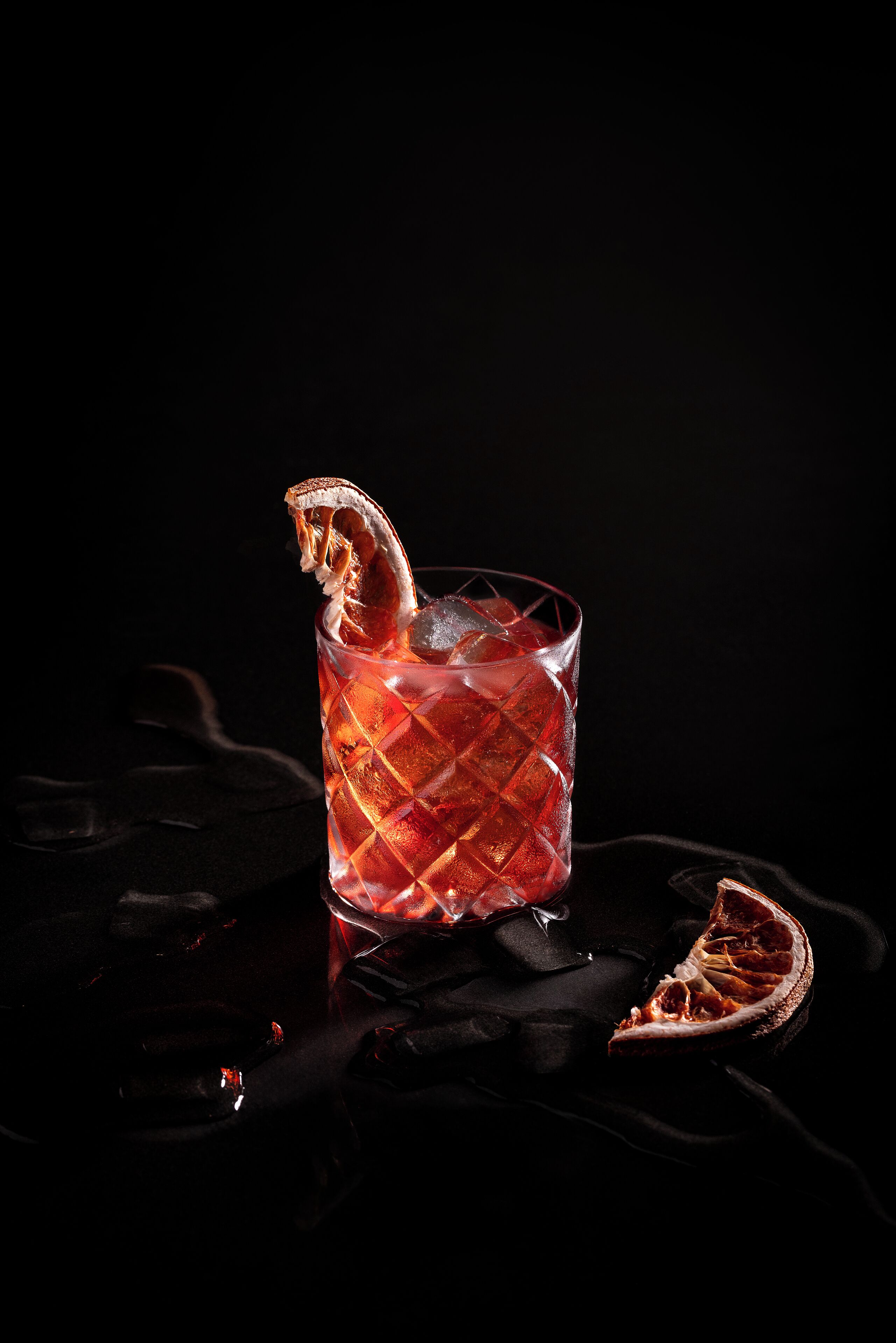 A glass of red beverage with a garnish, ice cubes, and a slice of citrus fruit on a reflective surface.