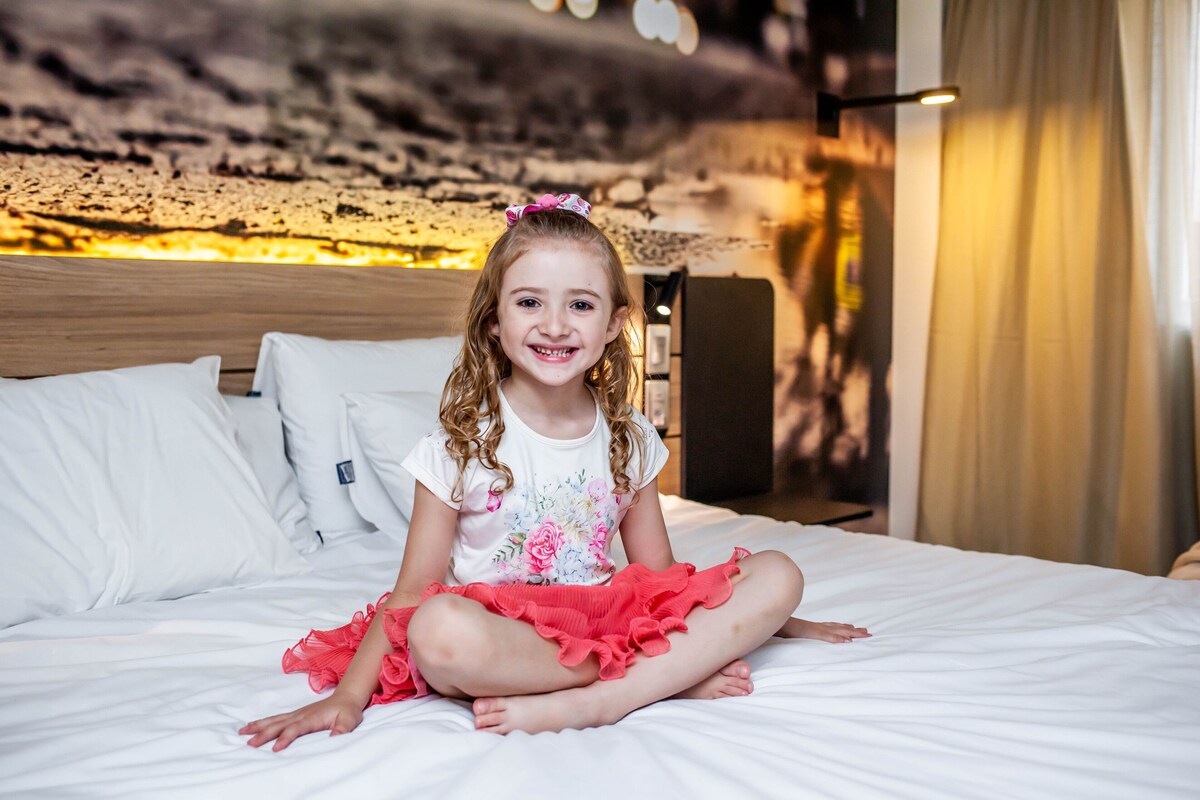 A child sitting on a bed in a hotel room with a wooden headboard and a wall with a textured design.