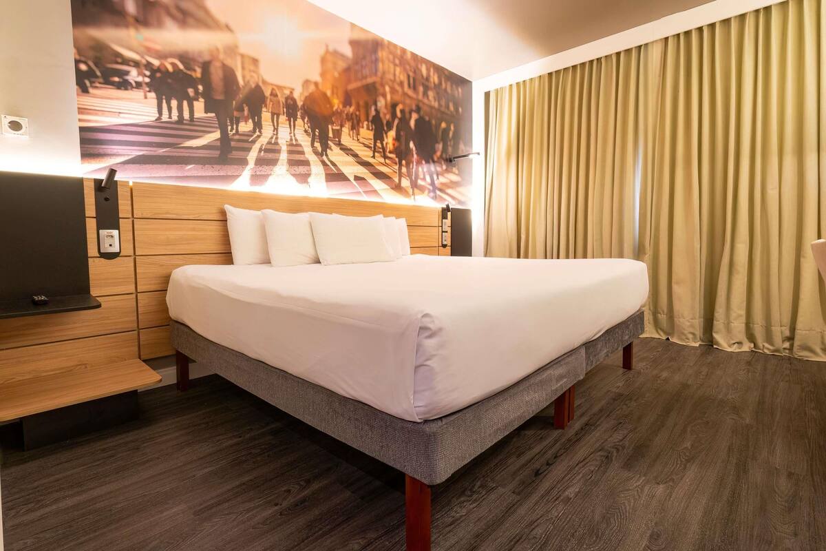 A hotel room with a large bed, wooden headboard, a flat-screen TV, and a cityscape mural on the wall.