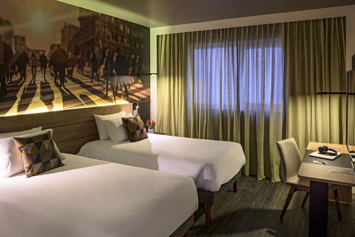 A hotel room with two beds, a desk, and a large window with curtains.