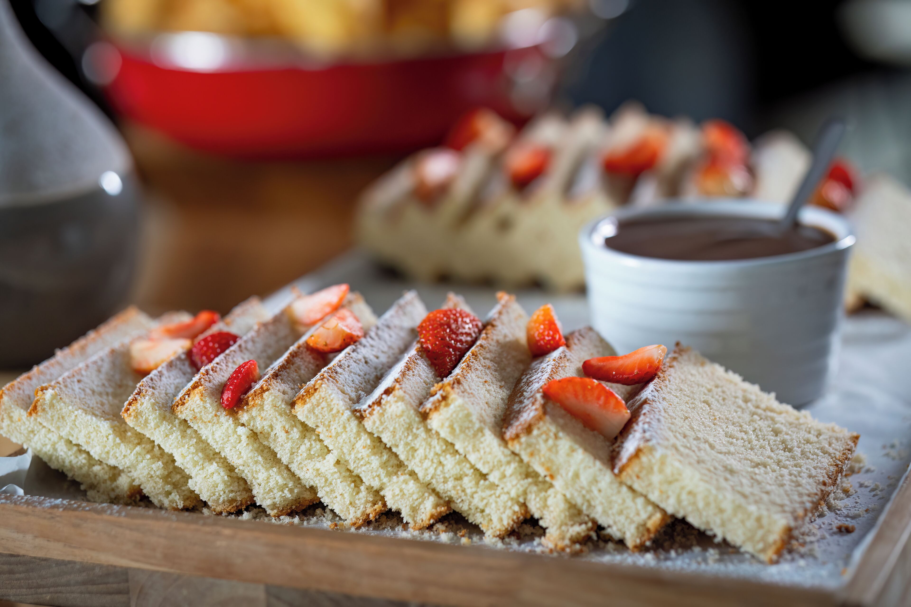A slice of cake with strawberries on top, dusted with powdered sugar, and a small cup of chocolate sauce.