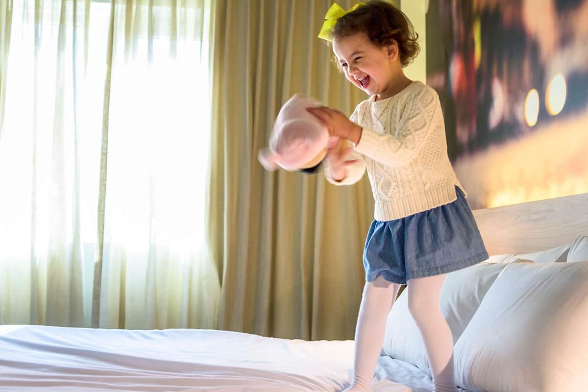 A child playing with a toy on a bed in a room with curtains and a bed with white bedding.