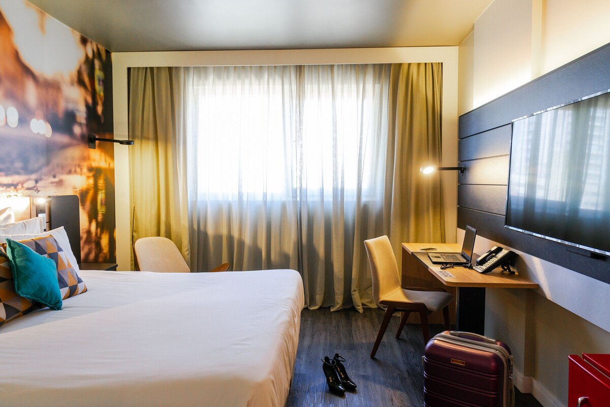 A hotel room with a bed, a desk with a laptop, a chair, a TV, and a suitcase.