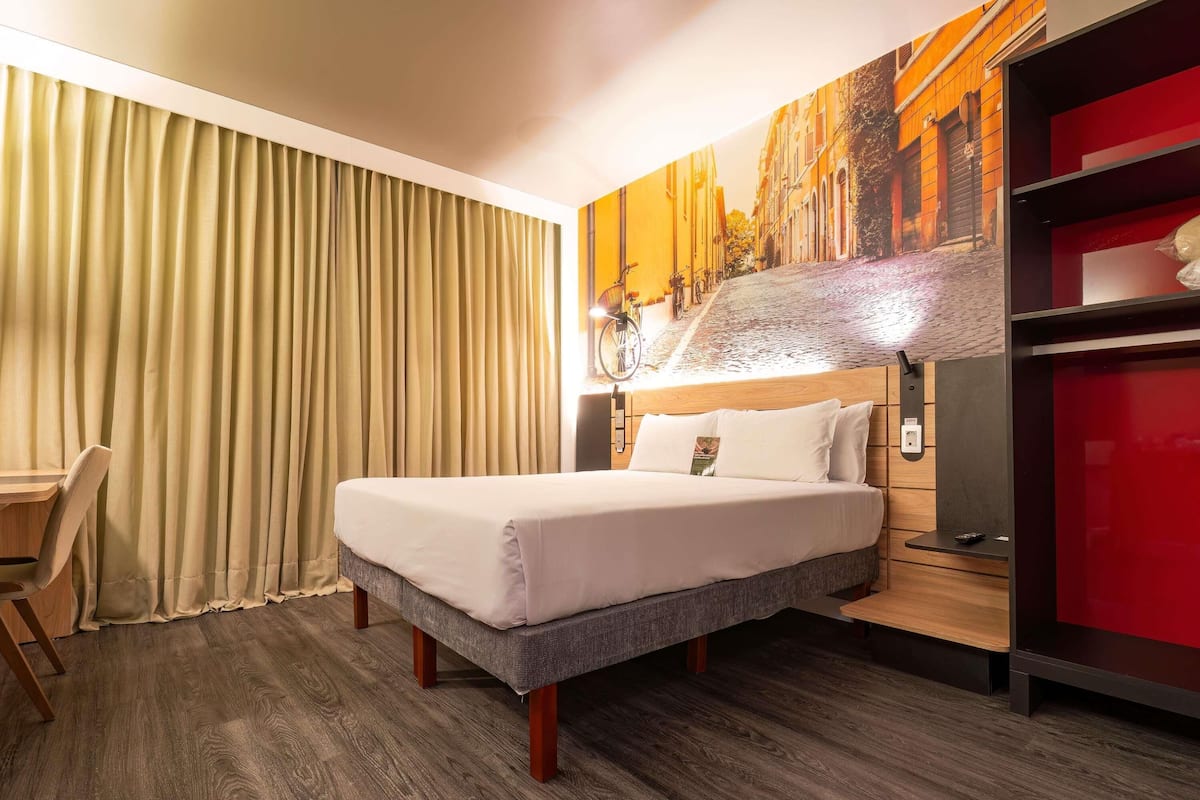 A hotel room with a bed, a desk, a chair, a painting on the wall, and a red cabinet.