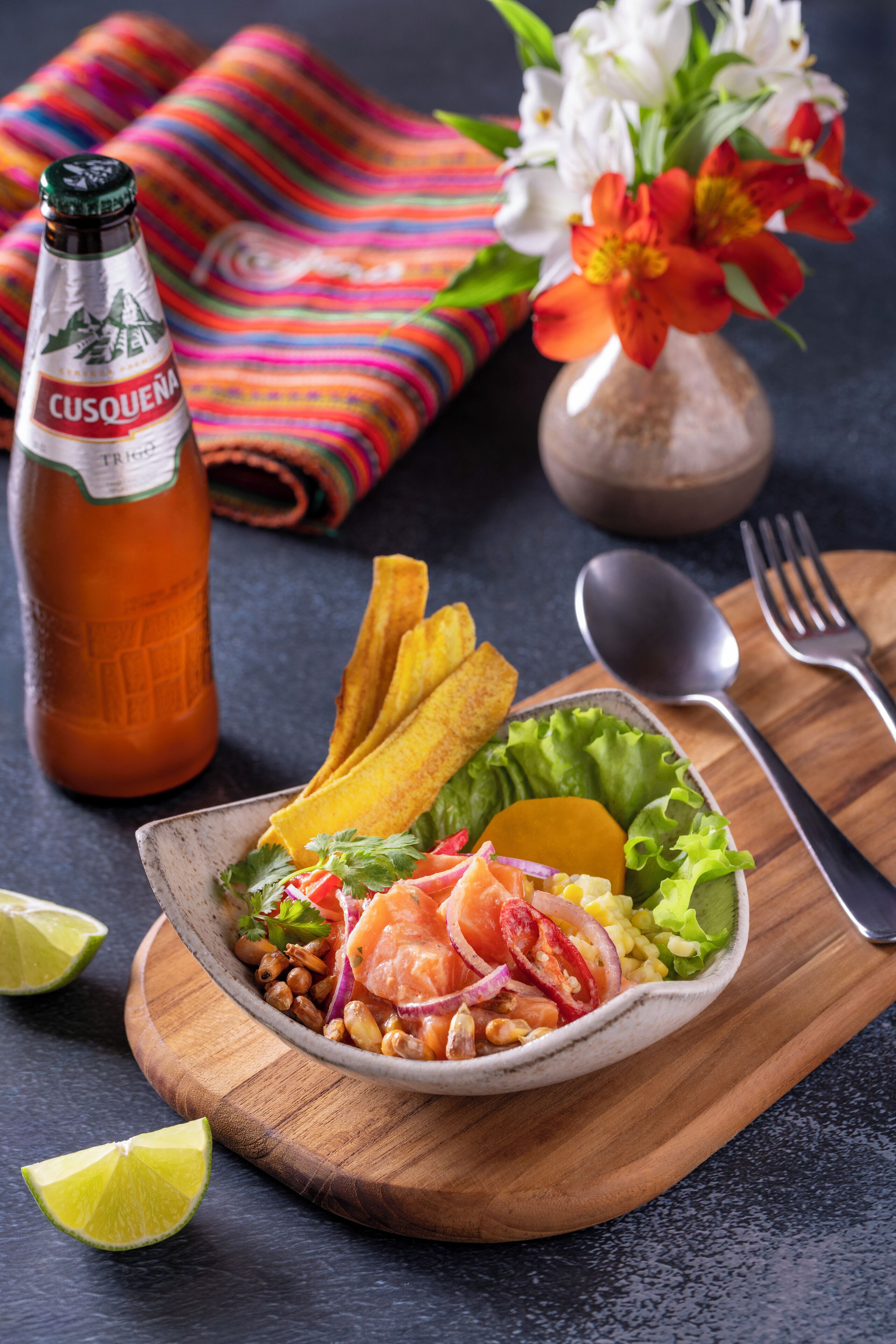 Un plato de comida con una botella de cerveza Cusquena, una cuchara, un tenedor y una servilleta sobre una tabla de madera.