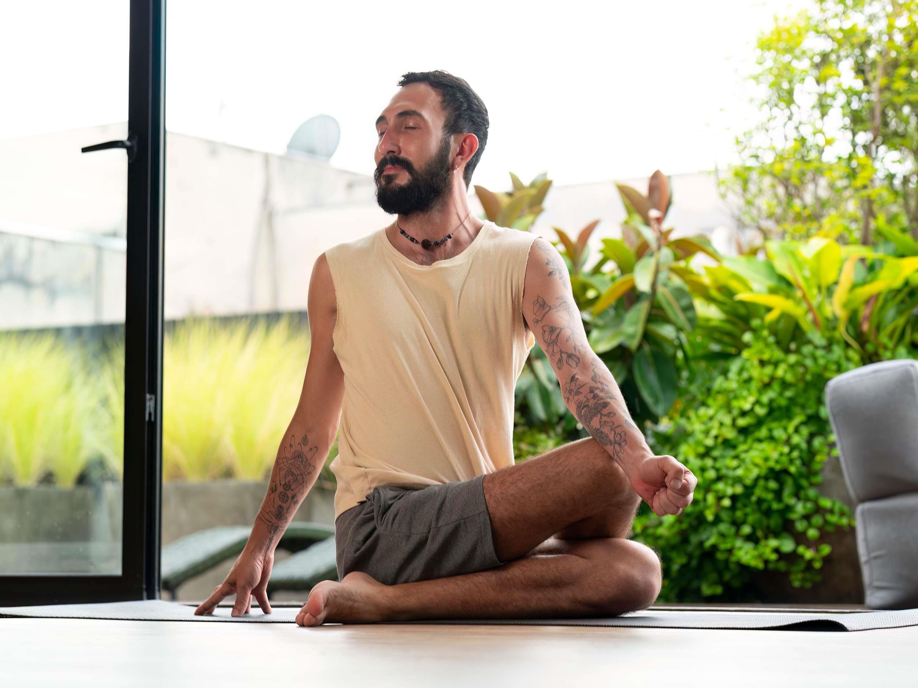Uma pessoa sentada sobre um tapete de yoga em ambiente interno, com vista para vegetação lá fora.