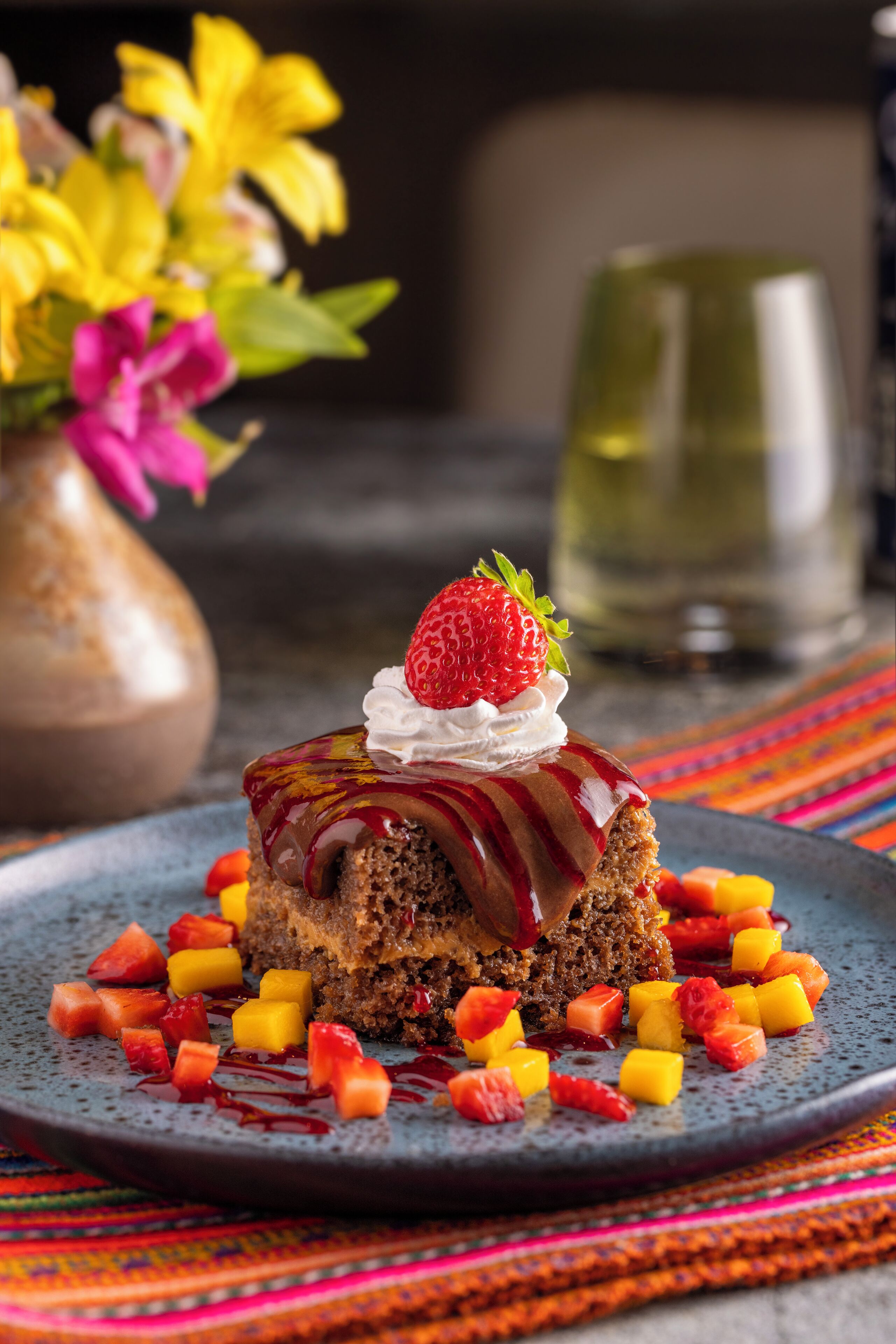 A dessert with strawberries and mango on a plate, with a vase of flowers and a glass in the background.
