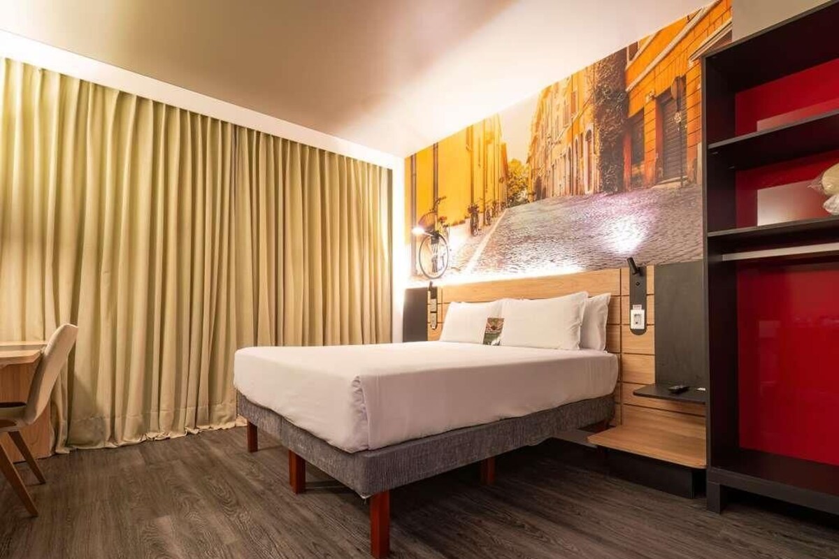A hotel room with a bed, a desk, a chair, a red cabinet, and a wall with a cityscape mural.