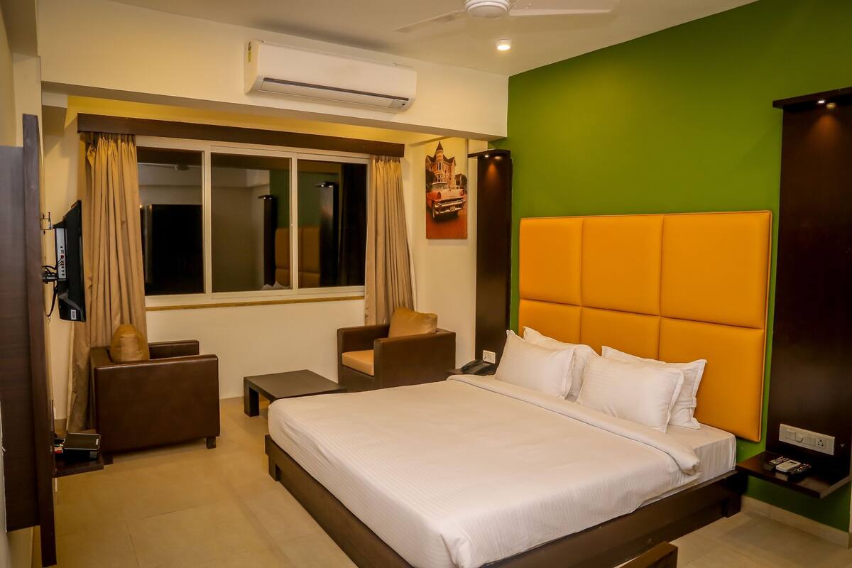 A hotel room with a large bed, a desk, a chair, a window with curtains, and an air conditioning unit.