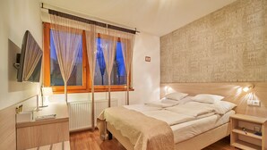 A bedroom with a bed, bedside tables, a lamp, a TV, and a window with curtains. - Oázis Apartmanok (Morahalom)