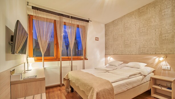 A bedroom with a bed, bedside tables, a lamp, a TV, and a window with curtains. - Oázis Apartmanok (Morahalom)